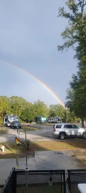 A rainbow arcs over an RV park with parked motorhomes, a white SUV, gravel sites, and surrounding trees after rain.