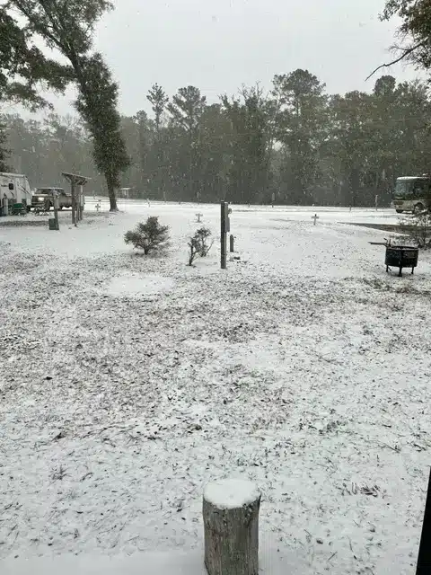 Snow falling over an RV park's open grassy sites with pine trees, a parked motorhome, utility posts, and a metal fire