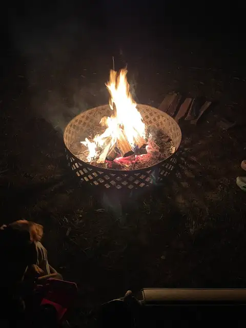 Nighttime campfire burning in a metal lattice fire ring with stacked firewood nearby and a child seated in foreground.