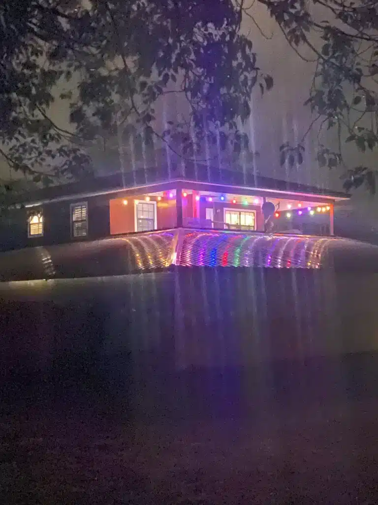 Nighttime photo of a single-story house with covered porch strung with colorful string lights and glowing lit windows