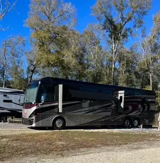 A large dark purple Class A motorhome parked on a gravel site with bare trees and blue sky behind.